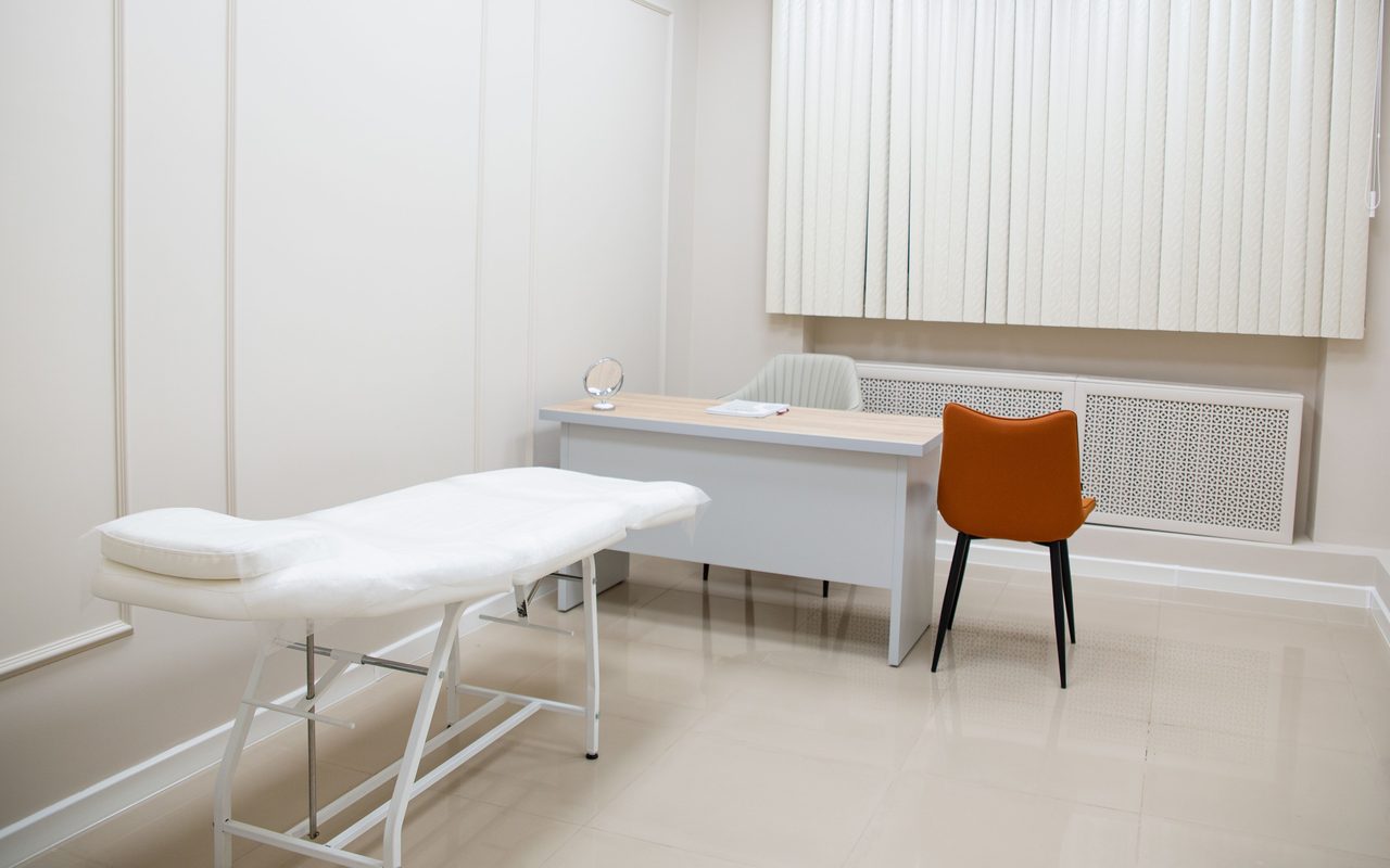 Minimal medical examination room with a bed, desk and chairs on a light tiled floor